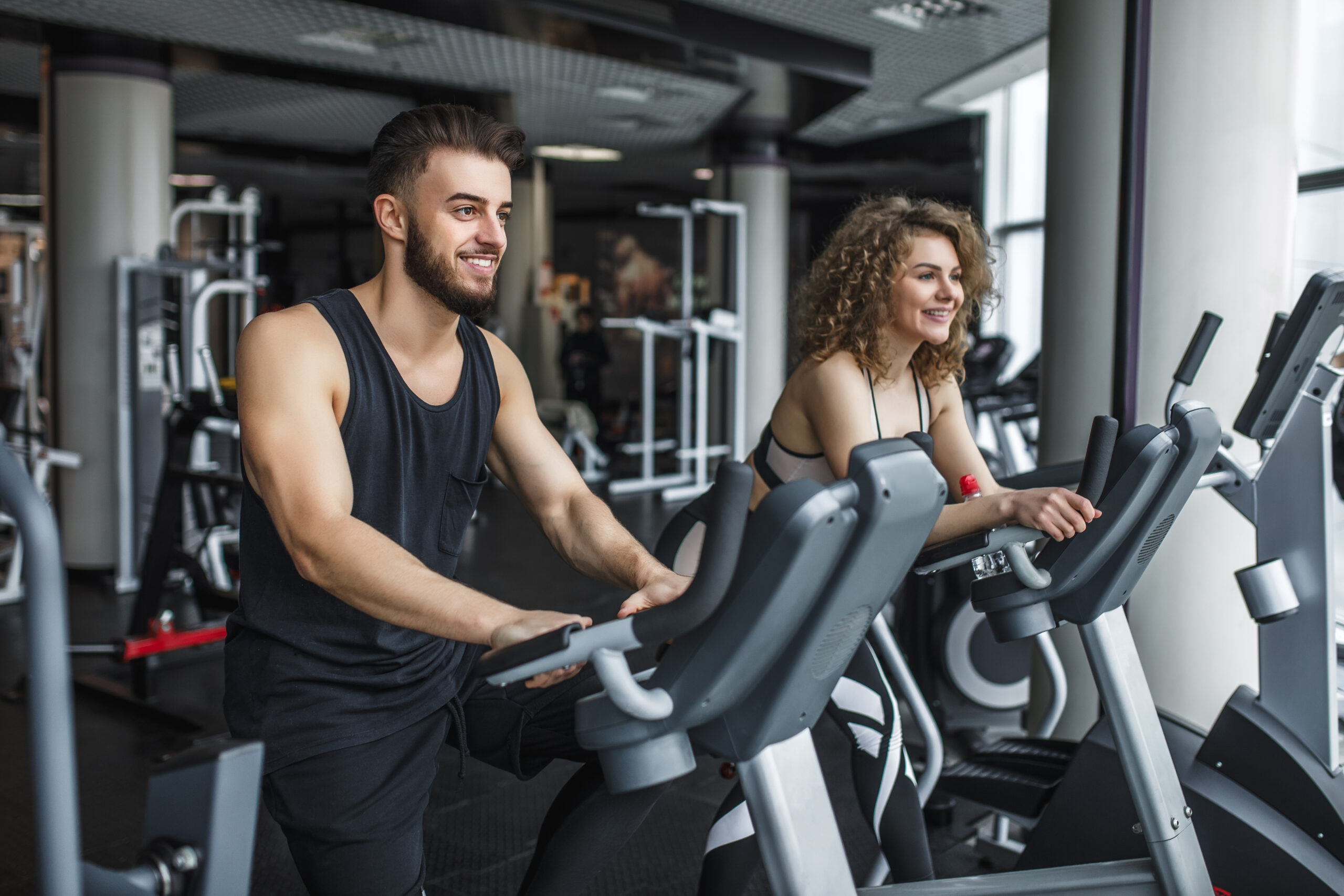 Attractive young woman and her trainer running on treadmill in gym. Slim girl jogging in fitness club, smiling at camera. Healthy lifestyle concept, cardio training.